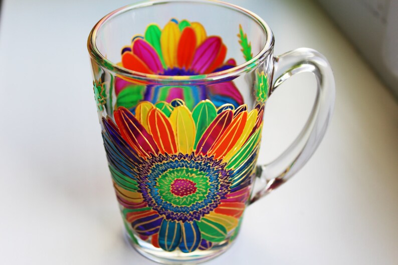 May include: Clear glass mug with a vibrant, hand-painted floral design. The colorful flower features petals in shades of red, orange, yellow, green, and purple, outlined in gold. The mug has a clear handle and is suitable for hot or cold beverages.