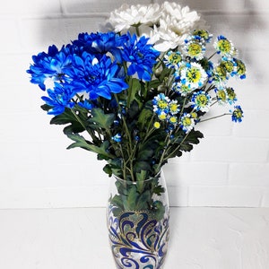 May include: A glass vase with a blue and gold decorative design holds a bouquet of blue, white, and yellow chrysanthemums. The flowers are arranged with green stems and leaves, creating a vibrant floral display against a white brick wall.