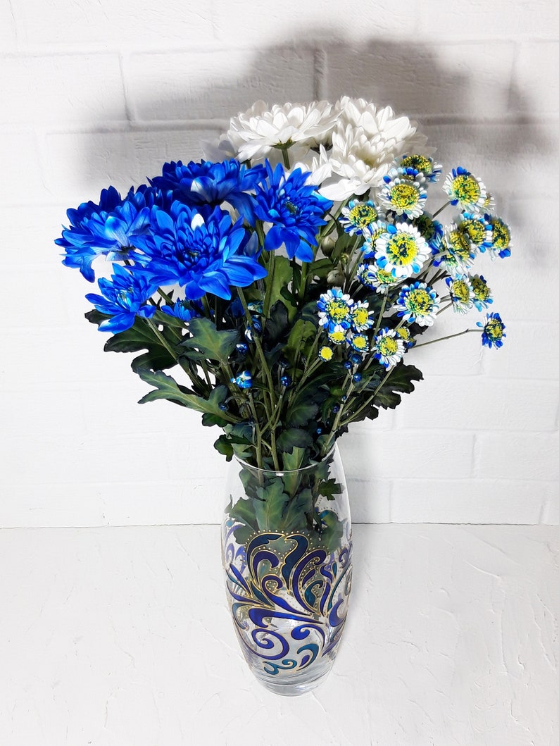 May include: A glass vase with a blue and gold decorative design holds a bouquet of blue, white, and yellow chrysanthemums. The flowers are arranged with green stems and leaves, creating a vibrant floral display against a white brick wall.
