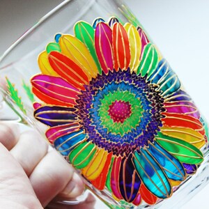May include: Clear glass mug with a hand-painted sunflower design. The sunflower features vibrant petals in shades of red, orange, yellow, green, blue, and purple, outlined in gold. The mug has a handle and is suitable for hot or cold beverages.