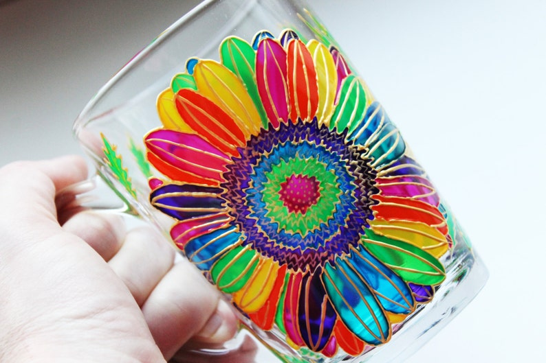 May include: Clear glass mug with a hand-painted sunflower design. The sunflower features vibrant petals in shades of red, orange, yellow, green, blue, and purple, outlined in gold. The mug has a handle and is suitable for hot or cold beverages.