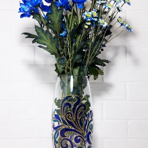 May include: A clear glass vase with a blue and gold decorative design, filled with blue, white, and yellow chrysanthemums. The vase has an oval shape and is approximately 10 inches tall. The flowers have green stems and leaves.