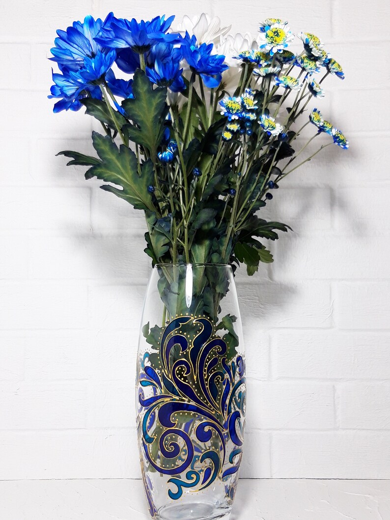 May include: A clear glass vase with a blue and gold decorative design, filled with blue, white, and yellow chrysanthemums. The vase has an oval shape and is approximately 10 inches tall. The flowers have green stems and leaves.