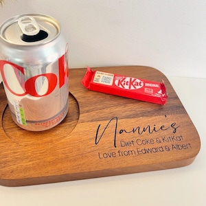 May include: A wooden tray with two circular cutouts for a can and a chocolate bar. The tray has the text "Nannie's Diet Coke & KitKat Love from Edward & Albert" engraved on it. A can of Diet Coke and a KitKat chocolate bar are placed in the cutouts.