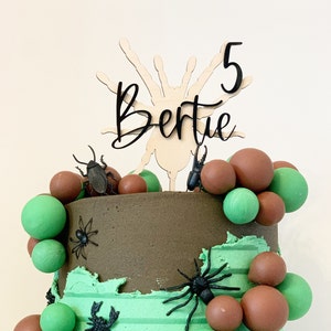 May include: A two-tiered chocolate cake decorated with green and brown balloons, black plastic spiders, and a wooden spider topper with the name "Bertie" and the number "5" in black lettering.