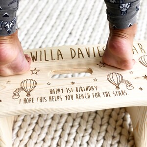 May include: A personalized wooden step stool with a hot air balloon and star design. The stool is engraved with the text "WILLA DAVIES" and "HAPPY 1ST BIRTHDAY I HOPE THIS HELPS YOU REACH FOR THE STARS."
