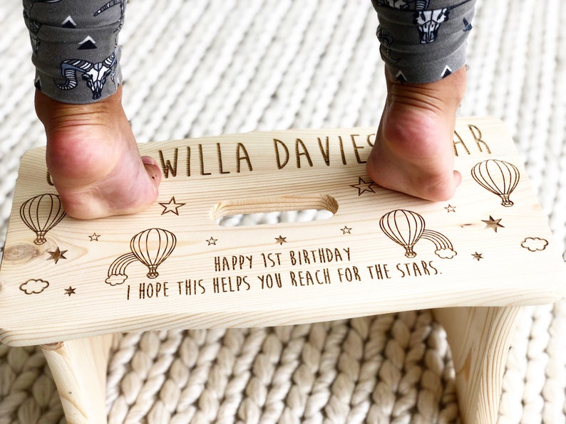 May include: A personalized wooden step stool with a hot air balloon and star design. The stool is engraved with the text "WILLA DAVIES" and "HAPPY 1ST BIRTHDAY I HOPE THIS HELPS YOU REACH FOR THE STARS."