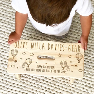 May include: A personalized wooden step stool with a light wood finish. The stool is decorated with hot air balloons, stars, and a rainbow. The text "OLIVE WILLA DAVIES-GEAR" is engraved on the top of the stool. The text "HAPPY 1ST BIRTHDAY I HOPE THIS HELPS YOU REACH FOR THE STARS." is engraved on the front of the stool.