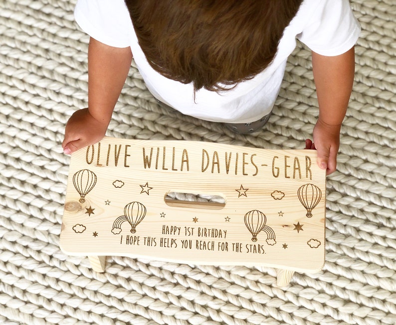 May include: A personalized wooden step stool with a light wood finish. The stool is decorated with hot air balloons, stars, and a rainbow. The text "OLIVE WILLA DAVIES-GEAR" is engraved on the top of the stool. The text "HAPPY 1ST BIRTHDAY I HOPE THIS HELPS YOU REACH FOR THE STARS." is engraved on the front of the stool.