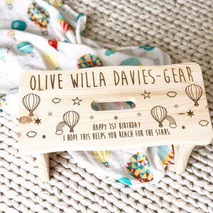 May include: A personalized wooden step stool with a light wood finish. The stool is decorated with a hot air balloon theme and the text "OLIVE WILLA DAVIES-GEAR HAPPY 1ST BIRTHDAY I HOPE THIS HELPS YOU REACH FOR THE STARS."