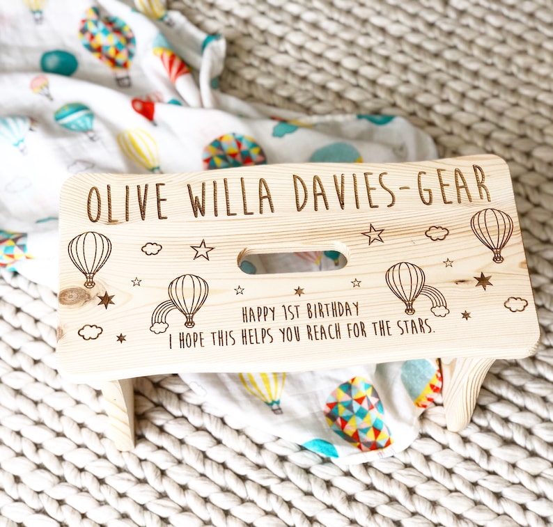 May include: A personalized wooden step stool with a light wood finish. The stool is decorated with a hot air balloon theme and the text "OLIVE WILLA DAVIES-GEAR HAPPY 1ST BIRTHDAY I HOPE THIS HELPS YOU REACH FOR THE STARS."
