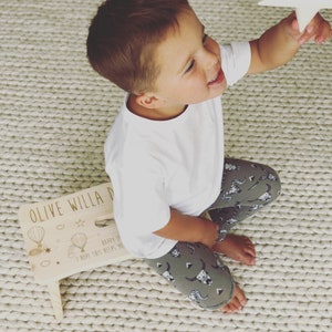 May include: A wooden step stool with the words "Olive Willa D" and "Happy 1st Birthday I hope this helps you reach for the stars" written on it. The stool is decorated with hot air balloons and stars. A young child is sitting on the floor next to the stool, reaching for a white star.
