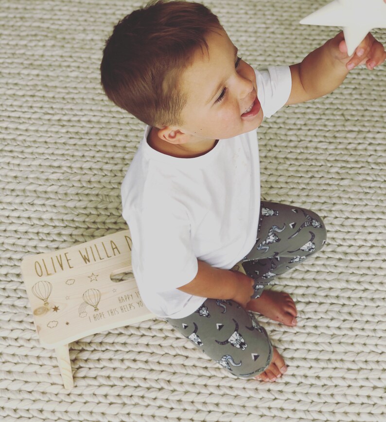May include: A wooden step stool with the words "Olive Willa D" and "Happy 1st Birthday I hope this helps you reach for the stars" written on it. The stool is decorated with hot air balloons and stars. A young child is sitting on the floor next to the stool, reaching for a white star.