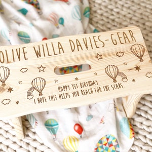 May include: A personalized wooden step stool with a hot air balloon theme. The stool is engraved with the name "Olive Willa Davies-Gear" and the message "Happy 1st Birthday. I hope this helps you reach for the stars."