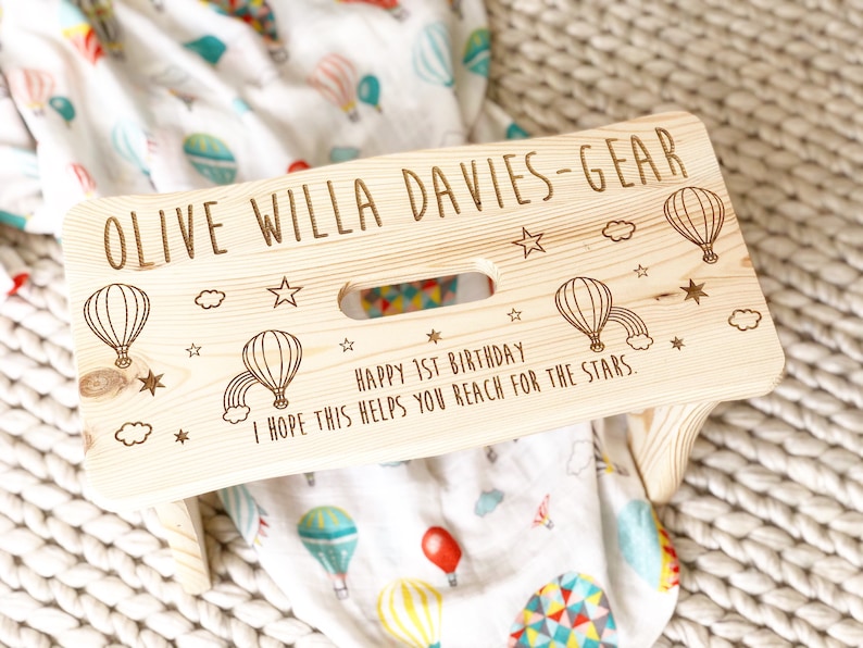 May include: A personalized wooden step stool with a hot air balloon theme. The stool is engraved with the name "Olive Willa Davies-Gear" and the message "Happy 1st Birthday. I hope this helps you reach for the stars."