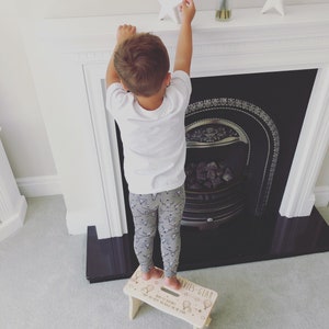 May include: A child standing on a wooden step stool with the text "Davies Gear" and "Happy 1st Birthday, Big Boy Keeps on Reaching for the Stars" printed on the top. The stool is decorated with a star, hot air balloons, and other small details.