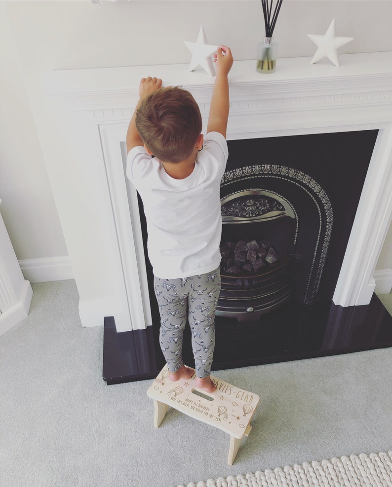 May include: A child standing on a wooden step stool with the text "Davies Gear" and "Happy 1st Birthday, Big Boy Keeps on Reaching for the Stars" printed on the top. The stool is decorated with a star, hot air balloons, and other small details.