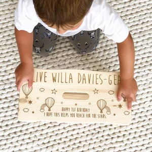 May include: A personalized wooden step stool with a hot air balloon theme. The stool is engraved with the text "LIVE WILLA DAVIES-GEA", "HAPPY 1ST BIRTHDAY", and "I HOPE THIS HELPS YOU REACH FOR THE STARS."