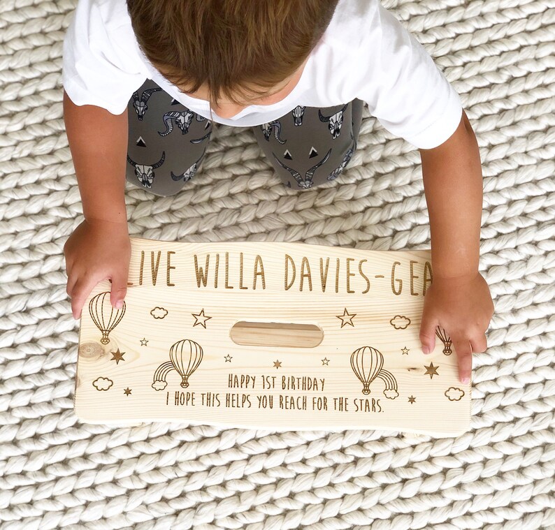 May include: A personalized wooden step stool with a hot air balloon theme. The stool is engraved with the text "LIVE WILLA DAVIES-GEA", "HAPPY 1ST BIRTHDAY", and "I HOPE THIS HELPS YOU REACH FOR THE STARS."