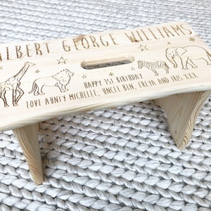 May include: A personalized wooden step stool with engraved animal illustrations of a giraffe, lion, zebra, and elephant. The stool has the name "ALBERT GEORGE WILLIAMS" and the text "HAPPY 1ST BIRTHDAY" engraved on it. It also includes a handle cutout.