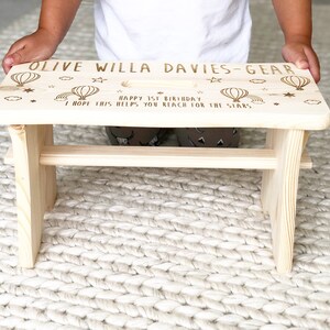 May include: A light wood step stool with a personalized message that reads "Olive Willa Davies-Gear Happy 1st Birthday I hope this helps you reach for the stars."