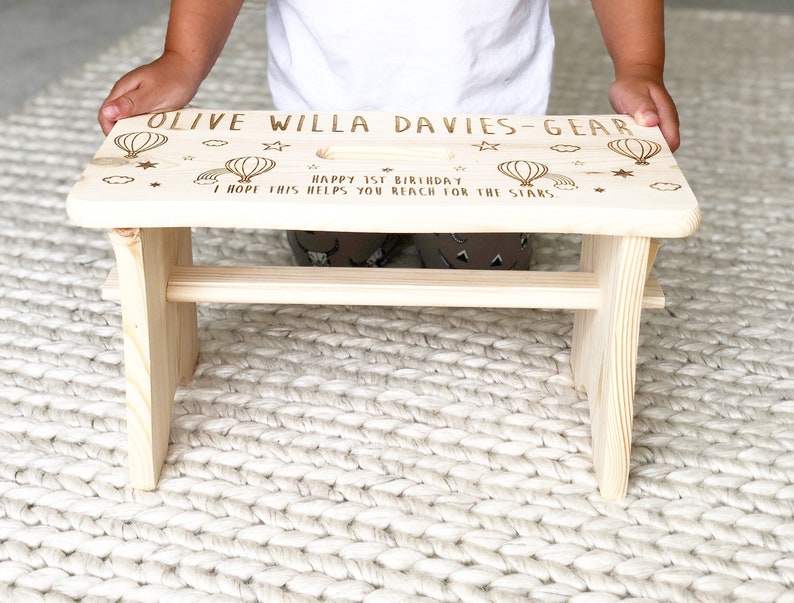 May include: A light wood step stool with a personalized message that reads "Olive Willa Davies-Gear Happy 1st Birthday I hope this helps you reach for the stars."