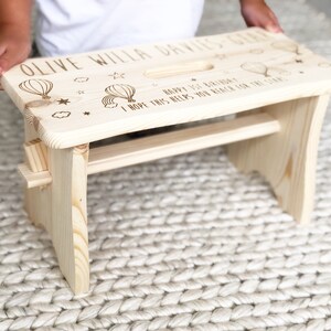 May include: A natural wood step stool with a personalized message that reads "Olive Willa Davies-Ger Happy 1st Birthday I hope this helps you reach for the stars". The stool has a small handle on the top and a cutout for a foot.