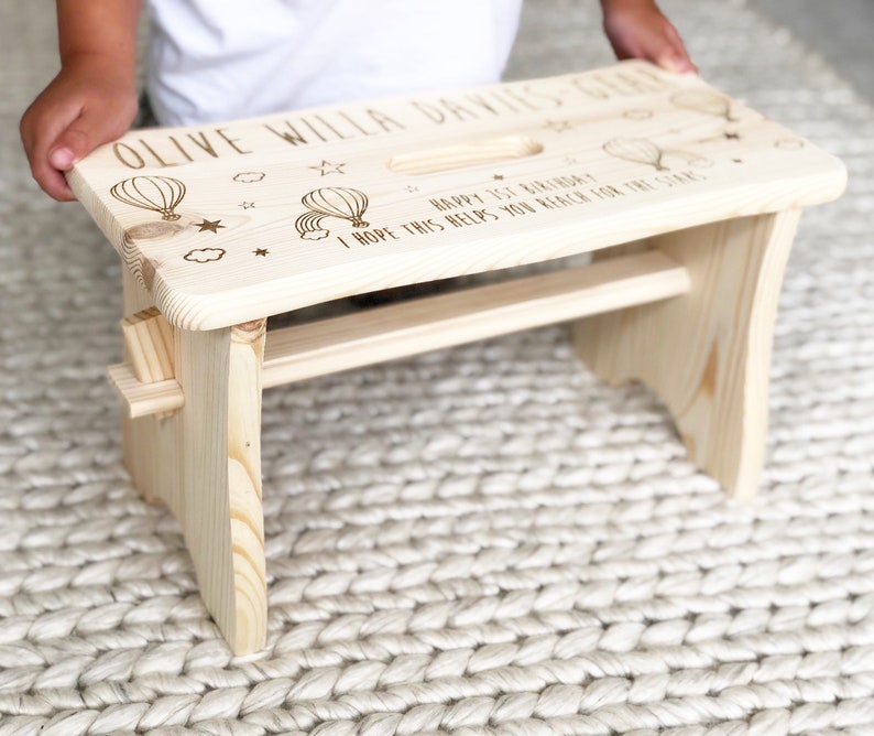 May include: A natural wood step stool with a personalized message that reads "Olive Willa Davies-Ger Happy 1st Birthday I hope this helps you reach for the stars". The stool has a small handle on the top and a cutout for a foot.