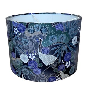 May include: Cylindrical lampshade with a Japanese-inspired design. The shade features a dark blue background with white cranes, blue flowers, and stylized wave patterns. The interior is white, and the overall aesthetic is elegant and decorative.