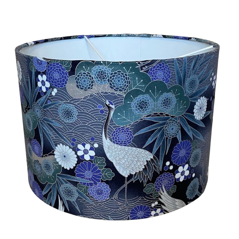 May include: Cylindrical lampshade with a Japanese-inspired design. The shade features a dark blue background with white cranes, blue flowers, and stylized wave patterns. The interior is white, and the overall aesthetic is elegant and decorative.