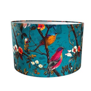 May include: Teal lampshade with a colourful bird and floral design. The lampshade features two birds perched on branches with white flowers and orange and yellow blooms. The background is teal.