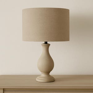 May include: A beige table lamp featuring a cylindrical lampshade and an urn-shaped base. The lampshade is a light tan colour, and the base is a lighter beige. The lamp is placed on a wooden surface.