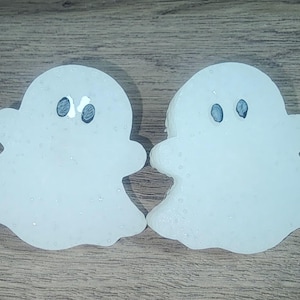 May include: Two white ghost-shaped cookies with black eyes. The cookies are decorated with white glitter.