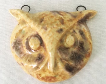 May include: A ceramic owl wall hanging with a speckled beige and brown finish. The owl has large, round eyes and a detailed beak. Two small, black metal loops are attached to the top for hanging. The owl's face is approximately 15 cm wide.