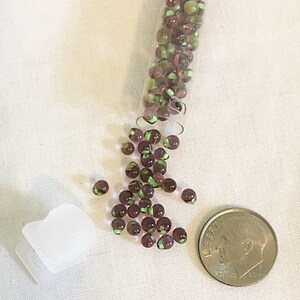 May include: A collection of small, round beads in shades of purple and green, spilling from a clear vial. A dime is next to the beads for size comparison. A small, white plastic container is also in the image.