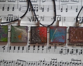 May include: Six square-shaped pendants with different abstract designs. The pendants are made of resin and have a silver-colored metal frame. The pendants are hanging on black cords.