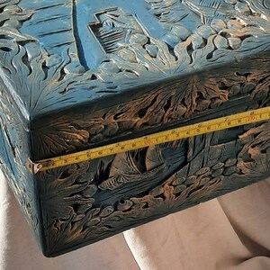 May include: A blue wooden box with intricate carvings of flowers and leaves. The box is closed and has a brass latch. A yellow measuring tape is placed along the edge of the box, showing a measurement of 11 inches.