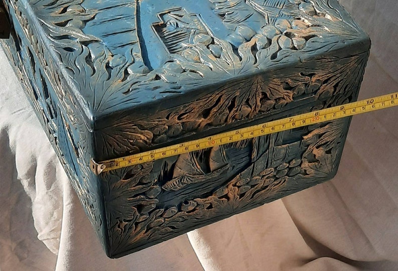 May include: A blue wooden box with intricate carvings of flowers and leaves. The box is closed and has a brass latch. A yellow measuring tape is placed along the edge of the box, showing a measurement of 11 inches.