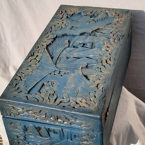 May include: A blue wooden box with intricate carvings of a landscape scene. The box has a hinged lid and is decorated with gold accents.