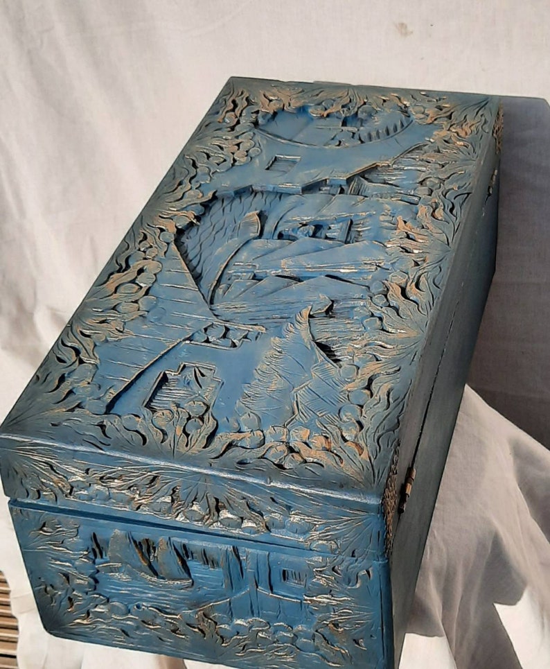 May include: A blue wooden box with intricate carvings of a landscape scene. The box has a hinged lid and is decorated with gold accents.