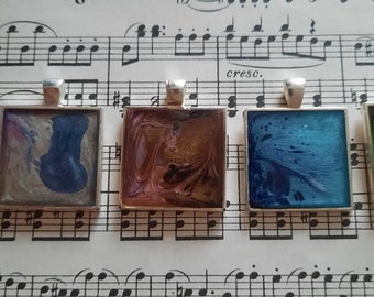 May include: Four square pendants with silver frames and colorful abstract resin art inside. The pendants are arranged in a row on a sheet of music.