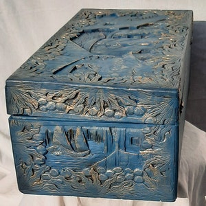 May include: A blue wooden box with intricate carvings of a floral design and a scene of boats on a lake. The box has a gold-colored paint finish that highlights the carvings.