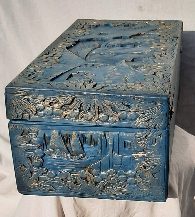 May include: A blue wooden box with intricate carvings of a floral design and a scene of boats on a lake. The box has a gold-colored paint finish that highlights the carvings.