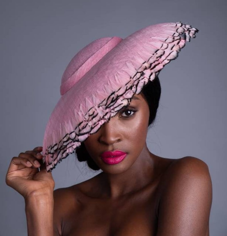 May include: A pink hat with a wide brim and a decorative feather trim. The hat is a light pink color and has a structured design. The brim is adorned with black and white feathers.