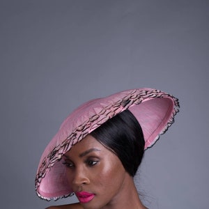 May include: A pink hat with a wide brim and a black feathered trim. The hat is angled, showcasing its design and texture. The hat is a fashion accessory.