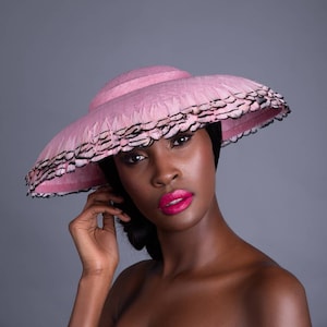 May include: A pink sun hat with a wide brim, trimmed with black and white feathers. The hat has a ribbon band and a structured crown. The hat is a stylish accessory, perfect for sun protection and fashion.