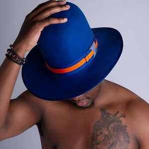 May include: A striking blue fedora hat featuring an orange and grey band. The hat has a wide brim and a tall crown. The hat is a stylish accessory. The person in the image is wearing a bracelet.