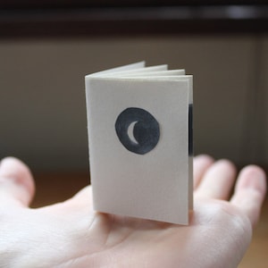 May include: A small, white book with a crescent moon cut out of the cover. The book is resting on a person's hand.