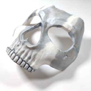 May include: A handcrafted skull mask in grey and white tones, featuring detailed teeth and eye sockets. The mask has a textured finish and is designed for wear. This unique mask is ideal for Halloween or costume parties.