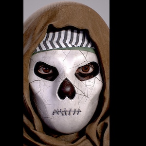 Peut inclure: Un masque de crâne blanc avec des orbites noires et une bouche cousue. Le masque est porté par une personne avec un vêtement à capuche marron et un bandana rayé noir et blanc.